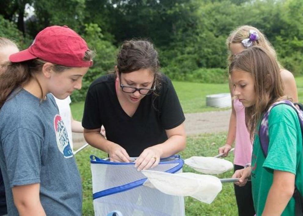 A person with glasses and brown hair who is a wearing a black shirt and who is teaching hands-on science to five girl scouts
