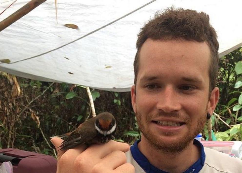 A person with brown hair who is in forested area of the Solomon Islands and who is holding a small, brown bird with a black and white face