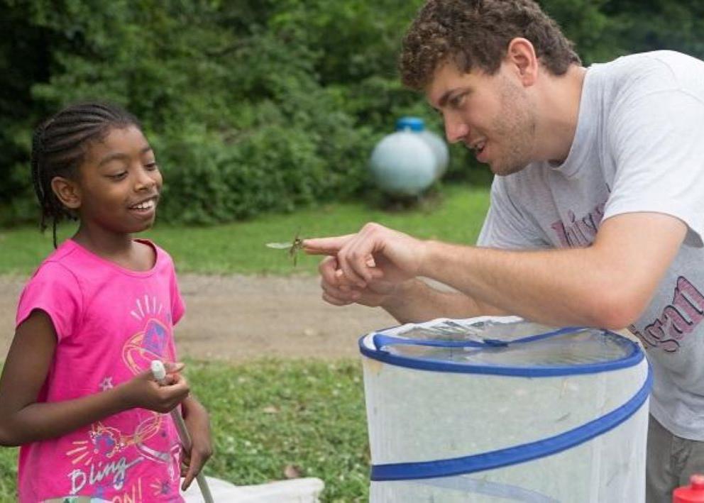 A person with curly brown hair who is teaching science to a girl scout who is wearing a pink shirt